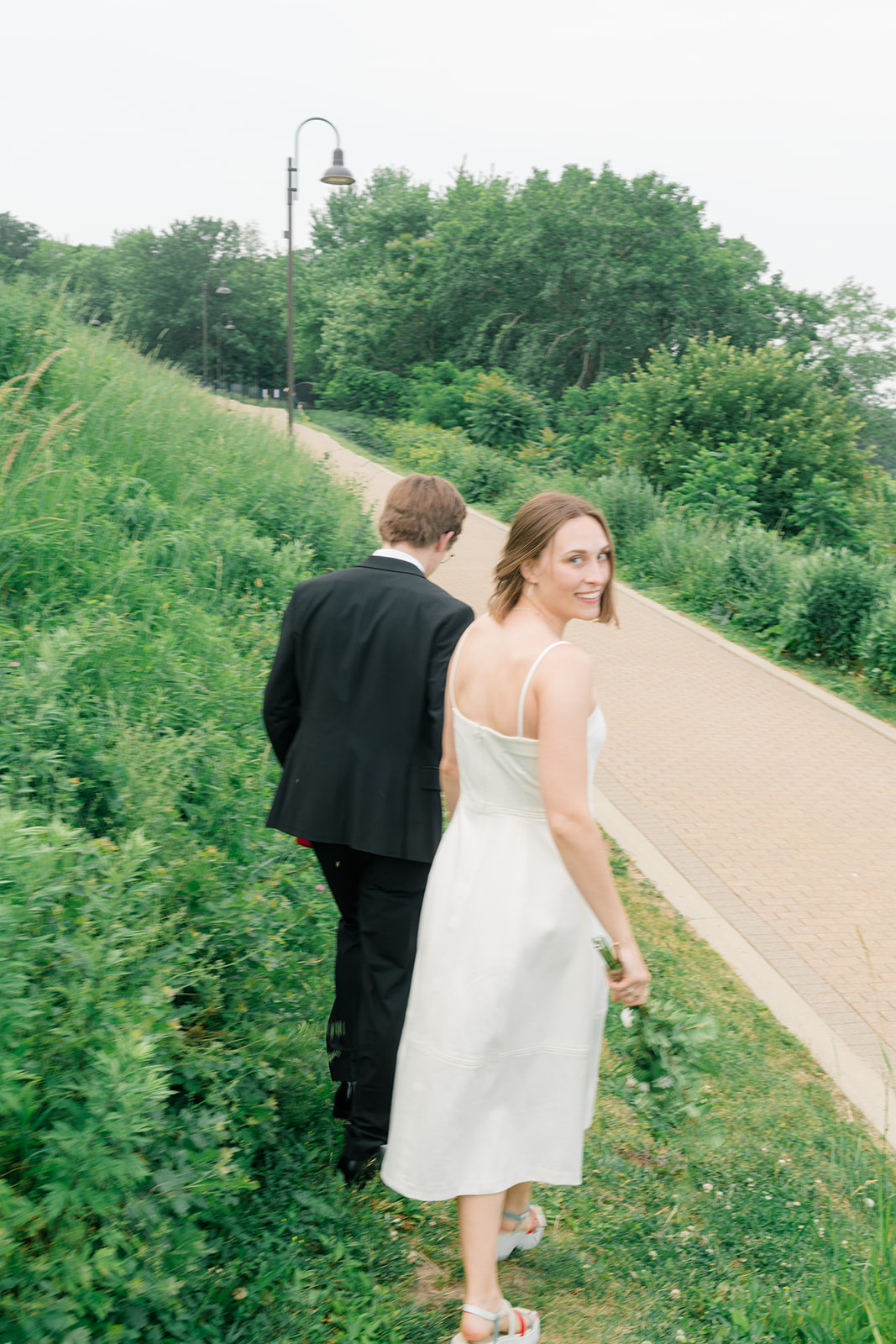Bride and groom walking together along a path at Lakewood Park in June after their Lakewood City Hall elopement, surrounded by lush greenery and soft summer light.